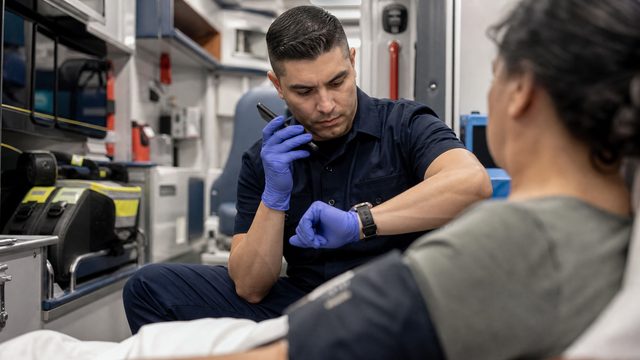 A paramedic checks his watch while waiting on a phone beside a patient in an ambulance